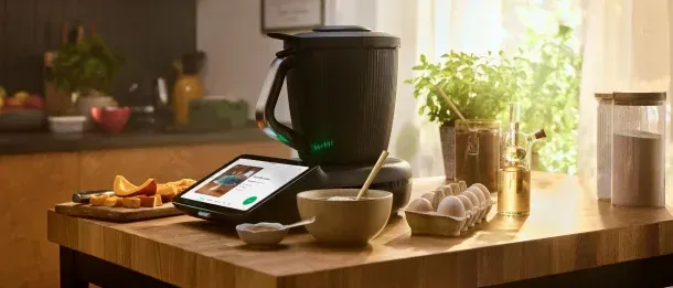Eine moderne Küche mit einem digitalen Display auf einem Küchengerät, einem Schneidebrett, Eiern und einer Topfpflanze. Das Gerät steht auf einem Holztisch.