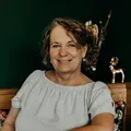 Eine Frau mit lockigem Haar lächelt und sitzt an einem Tisch mit einer schwarzen Schüssel und Tellern. Dahinter befindet sich ein Holzregal mit dekorativen Gegenständen.