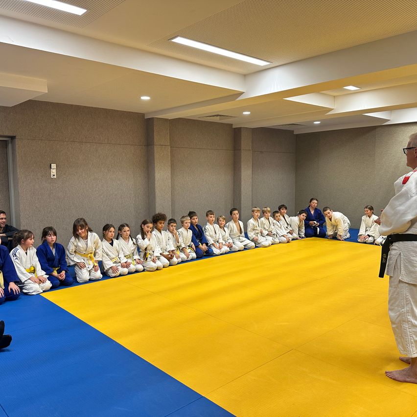 A group of young children dressed in blue and white judo uniforms are sitting on a blue and yellow mat, with a person in a white uniform standing at the front.