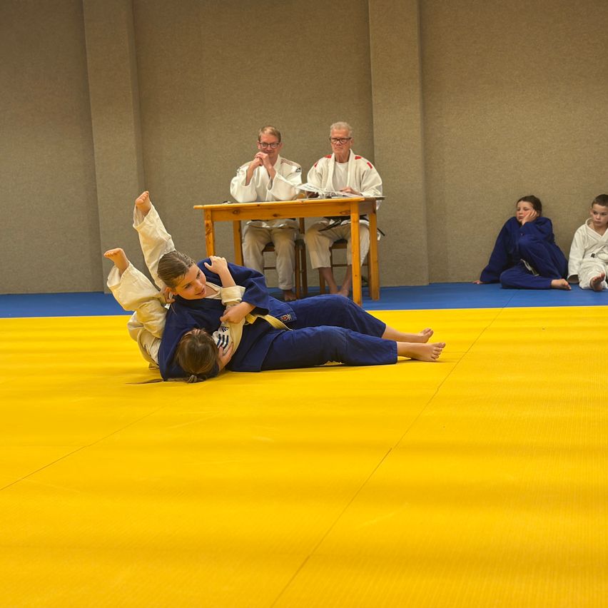 Two young boys in martial arts uniforms are grappling on a yellow mat, with two older men watching from a table.