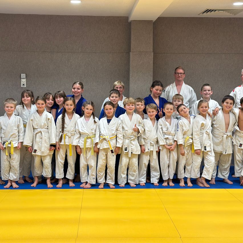 A group of children and young adults in martial arts uniforms stand in a line on a blue and yellow mat. An adult man and woman in martial arts uniforms are at the front.