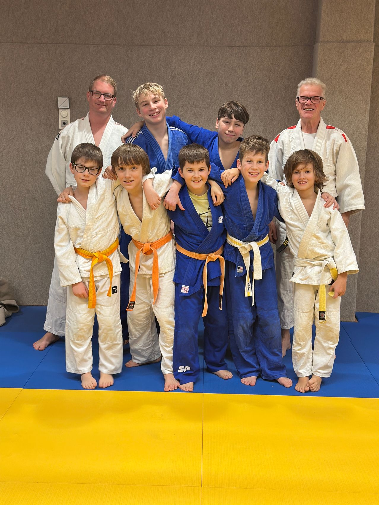 A group of young boys and two older men in martial arts uniforms pose for a photo on a blue mat. The boys wear white and orange belts, and the men wear white and red belts.