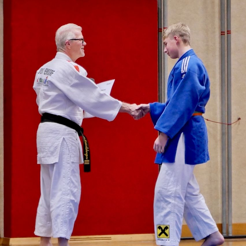 Ein Mann mit schwarzem Gürtel und weißem Gi schüttelt die Hand eines jungen Jungen in blauem Gi und orangem Gürtel in einem Kampfsportstudio.