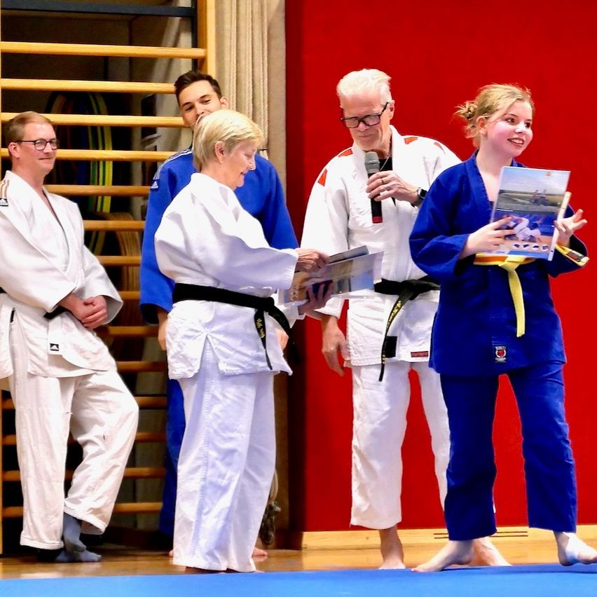 Eine Karate-Zeremonie mit Teilnehmern in blauen und weißen Uniformen. Ein Mann hält ein Mikrofon und spricht, während eine Frau ein Buch hält. Andere Teilnehmer beobachten.