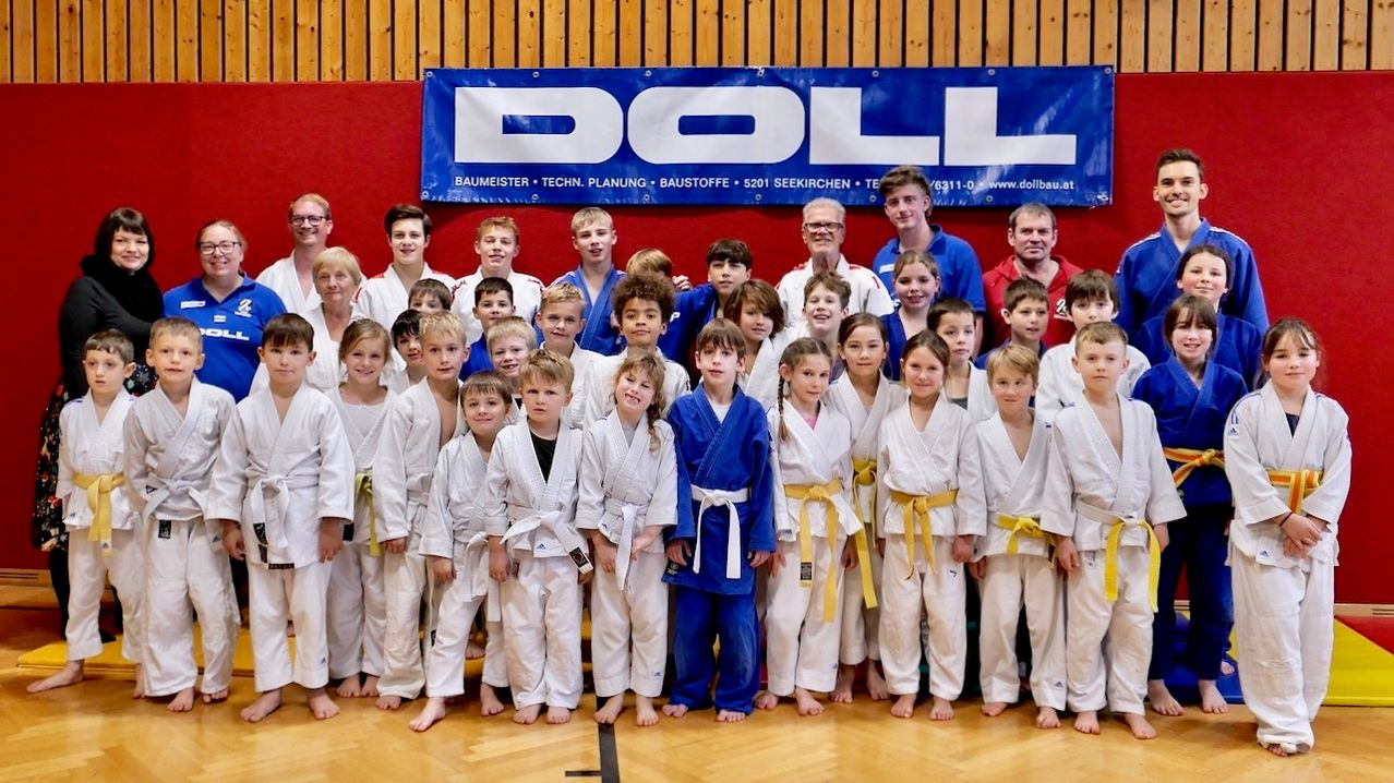 Eine Gruppe junger Kampfkünstler in weißen und blauen Uniformen posiert für ein Foto mit zwei Erwachsenen vor einem DOLL-Banner.