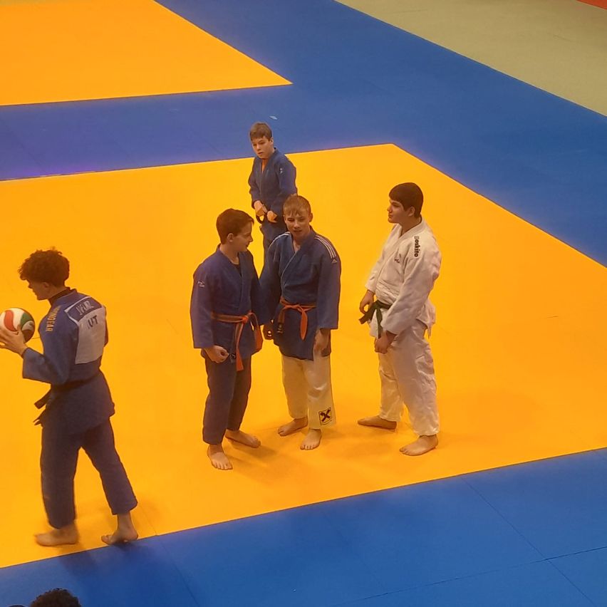 Fünf Jungen in Kampfsportuniformen stehen auf einem Matten. Einer hält einen Ball, während andere ihn ansehen. Sie sind barfuß, und die Matte ist gelb und blau.