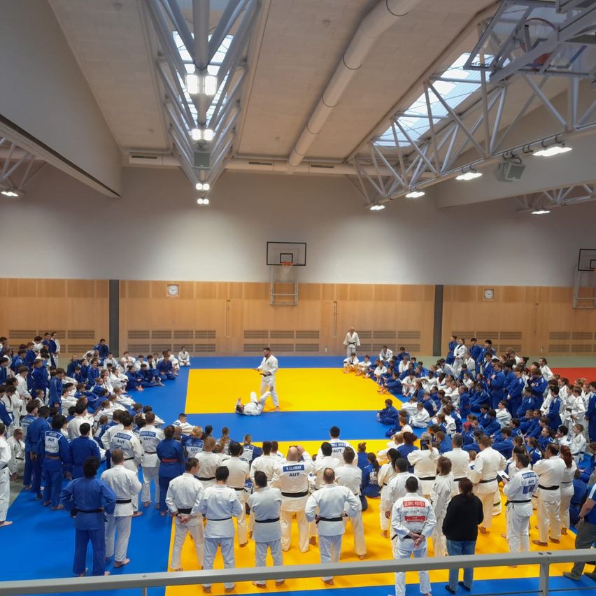 Eine Gruppe von Personen in blauen und weißen Uniformen ist um eine gelbe und blaue Matte versammelt, wahrscheinlich üben sie Kampfkunst. Eine Person ist auf der Matte, während andere in einem Kreis stehen. Es gibt Metallkonstruktionen darüber und einen Basketballkorb an der Wand.