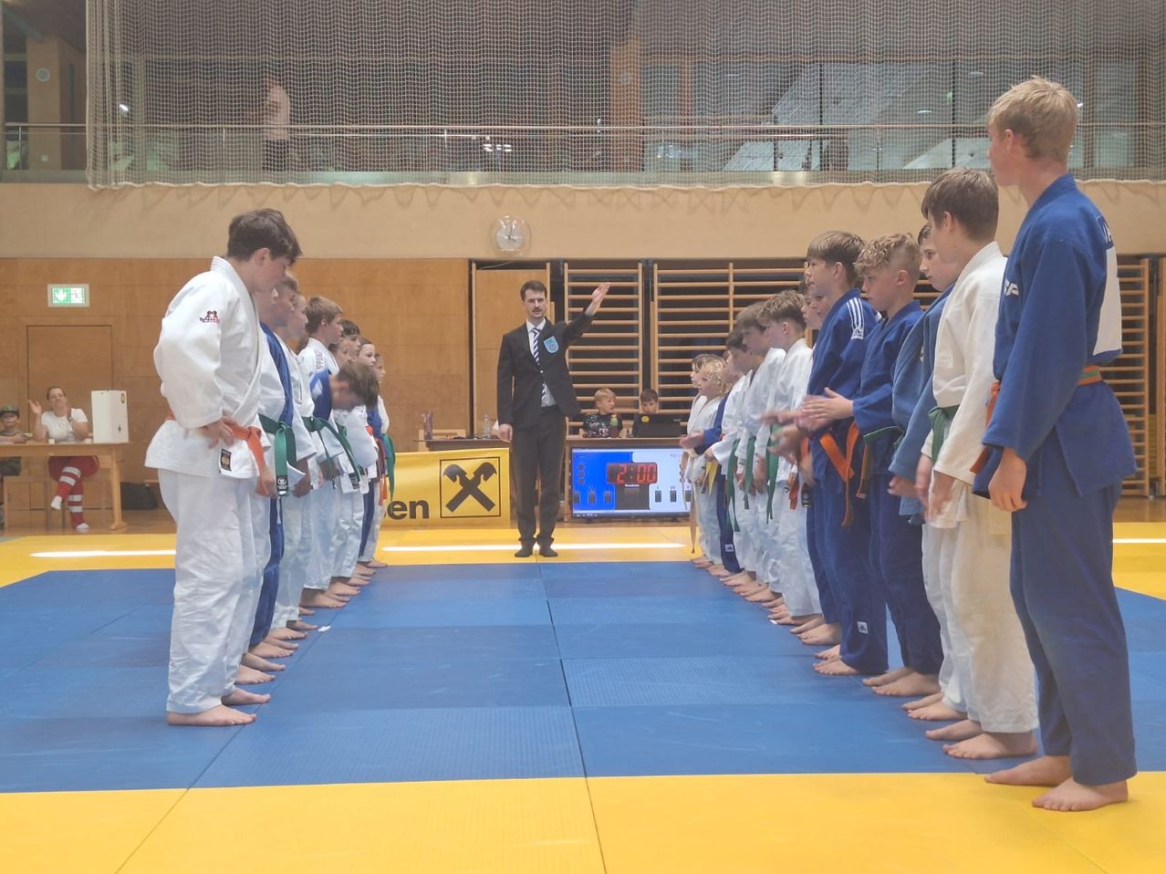 Eine Gruppe junger Jungen in Kampfsportuniformen steht auf einer Matte mit einem Schiedsrichter in der Mitte. Im Hintergrund ist ein Banner sichtbar.