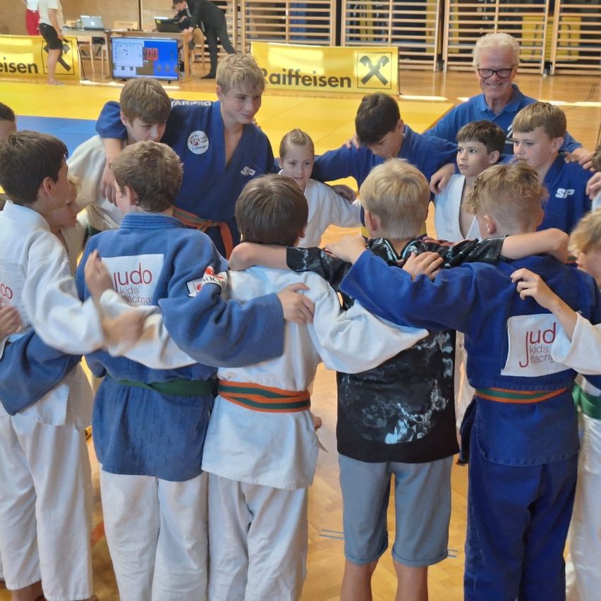 Kinder in Judo-Uniformen, einschließlich Kids, stehen in einem Kreis mit Erwachsenen, einer trägt eine Brille, in einer Sporthalle mit einem gelben Banner.