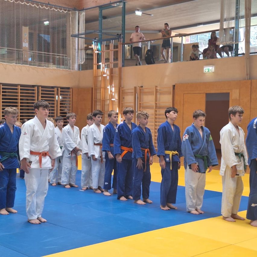 Eine Gruppe junger Männer in Judo-Uniformen steht in einer Reihe auf einem blauen Mattenboden. Zuschauer beobachten von der Galerie aus. Der Ort scheint ein Judo-Dojo zu sein.