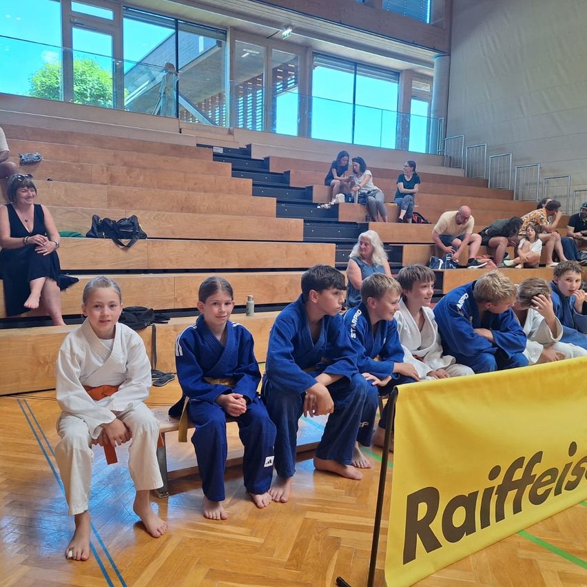 Mehrere Kinder in Kampfsportuniformen sitzen auf einer Bank vor einem Raiffeisen-Banner. Dahinter befinden sich Holztribünen mit sitzenden Personen. Die Szene spielt in einer Turnhalle.