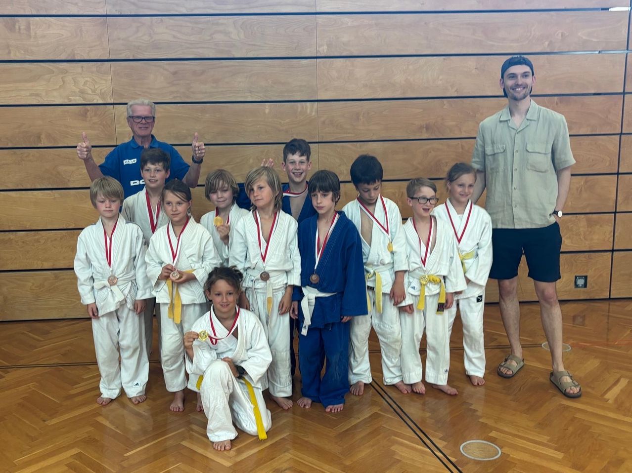 Eine Gruppe junger Karate-Schüler mit Medaillen steht mit ihren Trainern auf einem Holzboden. Die Schüler tragen weiße Uniformen, einige tragen Blau. Die Trainer sind in Freizeitkleidung.