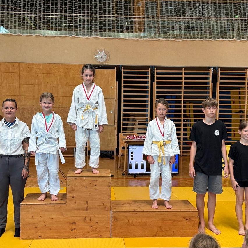 Eine Gruppe junger Judokas steht auf einer Holzplattform und trägt Medaillen und Uniformen. Ein Mann steht links und ein Junge rechts. Dahinter befindet sich ein gelber Teppich, eine Holzwand und eine Uhr.