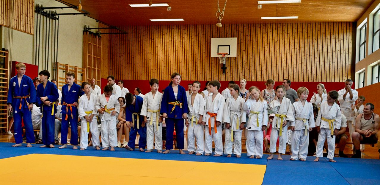 Eine Gruppe junger Kampfsportler in weißen und blauen Uniformen steht in einer Reihe auf einer Matte. Eine Frau in einer blauen Uniform steht vorne.