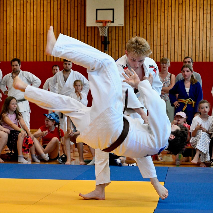 Ein Judo-Match findet in einer Turnhalle statt. Zwei junge Männer führen einen Judo-Wurf durch, mit Zuschauern, die zuschauen.