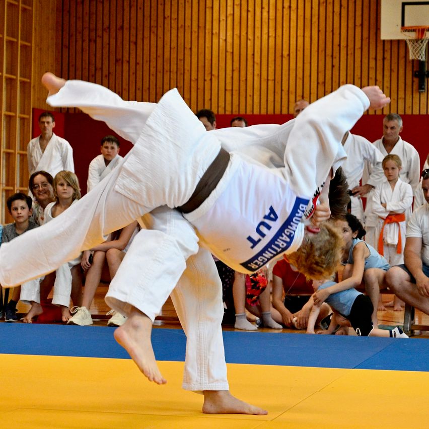 Ein Judo-Kampf ist im Gange. Ein Wettbewerber in Weiß hat den Gegner in einer Haltung. Zuschauer beobachten von den Seitenlinien.