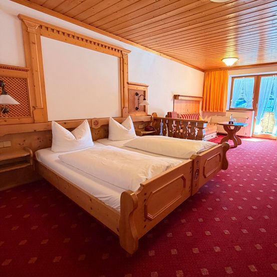 Ein gemütliches Schlafzimmer mit Holzbett, weißen Kissen und rotem Teppich. Holzwände, Decke und Möbel. Zwei Fenster mit orangefarbenen Vorhängen.