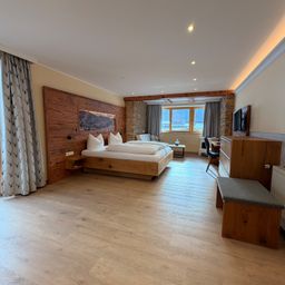 Ein Schlafzimmer mit Holzwänden und -böden, einem Doppelbett und einem Schreibtisch mit Stuhl. Es gibt ein Fenster mit Vorhängen, ein Bilderrahmen an der Wand und einen Heizkörper am Boden.