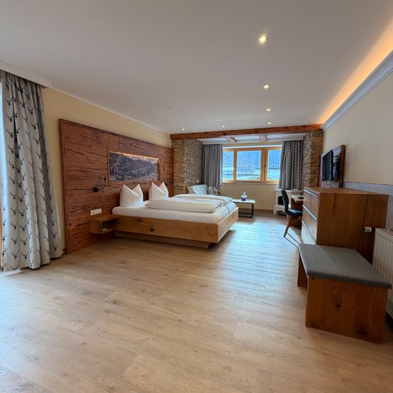 Ein Schlafzimmer mit Holzwänden und -böden, einem Doppelbett und einem Schreibtisch mit Stuhl. Es gibt ein Fenster mit Vorhängen, ein Bilderrahmen an der Wand und einen Heizkörper am Boden.