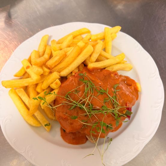 Ein Teller mit einem Fleischgericht in Tomatensauce mit Kräutergarnitur, begleitet von einer großzügigen Portion Pommes Frites.