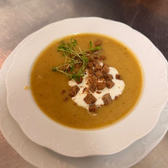 Ein weißer Teller mit goldgelber Suppe, gekrönt von Schlagsahne, Croutons und einem grünen Zweig. Es steht auf einem weißen Teller auf einer metallischen Oberfläche.