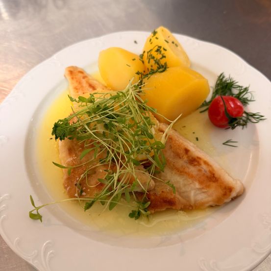 Ein weißer Teller mit gebratenem Fisch, gelben Kartoffeln und einer kleinen roten Tomate auf der Seite, garniert mit grünen Kräutern und serviert mit einer gelben Sauce.