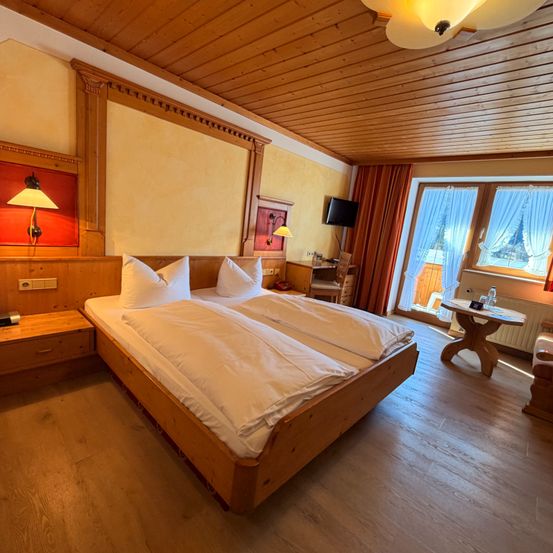 Ein gemütliches Schlafzimmer mit einem Doppelbett, einem Holzkopfteil und an der Wand montierten Lampen. Ein Schreibtisch, ein Stuhl und ein Fernseher stehen in der Ecke. Zwei Fenster mit Vorhängen bieten einen Blick nach draußen.