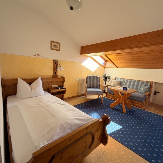 Ein Schlafzimmer mit einem Holzbett, weißen Bettlaken und Kissen. Ein offenes Fenster und eine kleine Couch sind im Zimmer.