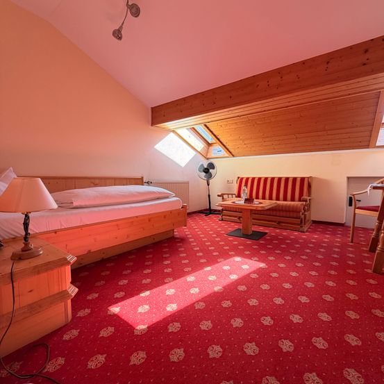 Ein gemütliches Schlafzimmer mit einem Holzbett, einer Lampe und einer Couch. An der Wand steht ein Ventilator und ein Heizkörper. Ein hölzerner Tisch steht in der Nähe der Couch. Der Raum hat eine Holzdecke und einen Teppichboden.