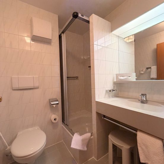 Ein modernes Badezimmer mit weißen Fliesen, einer Dusche und einem Waschbecken mit Spiegel. Ein Hocker und ein Handtuch sind in der Nähe.