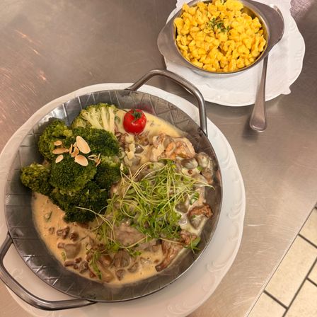 Eine Metallpfanne mit cremigem Pilzgericht, Brokkoli und Kräutern. Eine kleine Schüssel mit gelben Nudeln und eine Tomate auf einem weißen Teller.