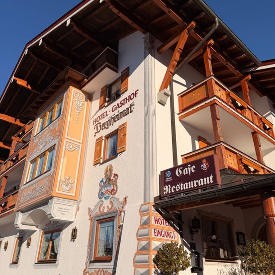 Das Hotel Gasthof Bergheimat hat ein weißes Äußeres mit braunen Verzierungen. Das Gebäude hat mehrere Fenster und Balkone mit dekorativen Verkleidungen. Das Café-Schild ist rot und weiß.