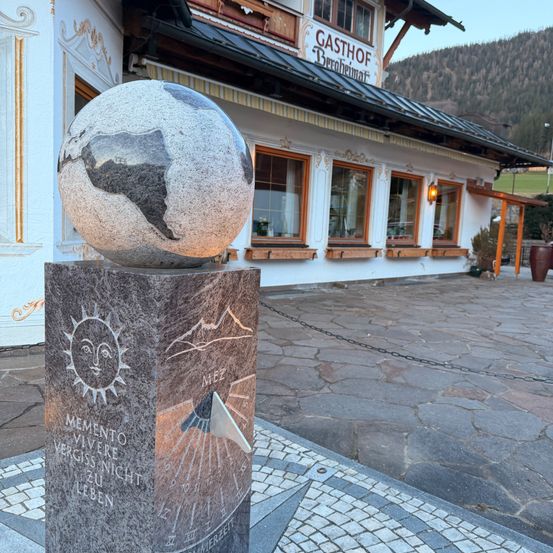 Ein kugelförmiges Denkmal steht vor einem Gebäude mit der Aufschrift Gasthof Berhheimat. Es hat einen Kompass und eine Sonnenuhr mit einer Sonne und einem Bergdesign.