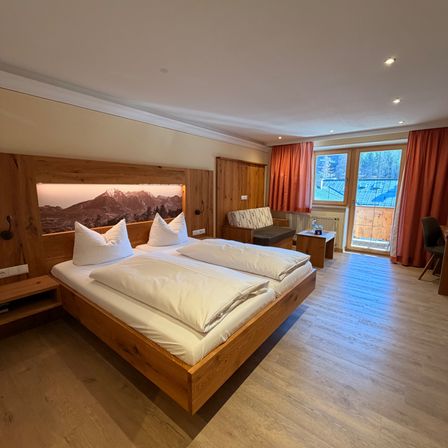 Ein gemütliches Schlafzimmer mit einem Doppelbett, einem holzverkleideten Kopfteil, weißen Laken und einem Holzstuhl in der Nähe einer Glastür mit Vorhängen.