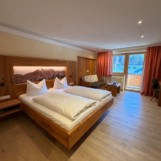 Ein gemütliches Schlafzimmer mit einem Doppelbett, einem holzverkleideten Kopfteil, weißen Laken und einem Holzstuhl in der Nähe einer Glastür mit Vorhängen.
