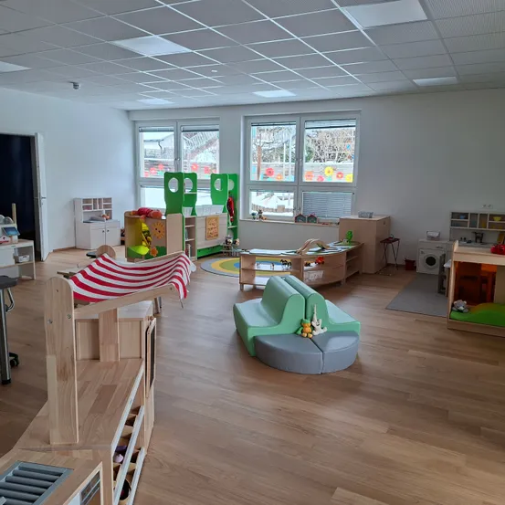 Ein gut beleuchteter Indoor-Spielbereich mit Holzmöbeln, bunten Spielzeugen und großen Fenstern mit Blick nach draußen.
