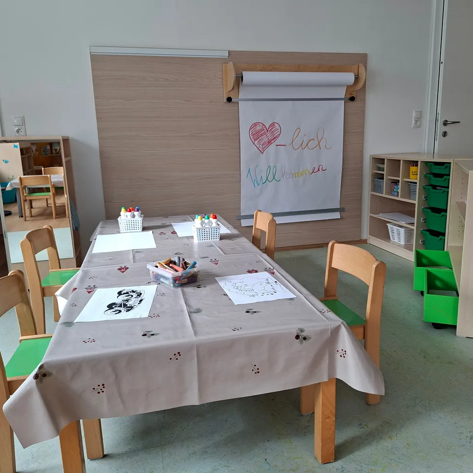Ein Klassenzimmer mit einem Tisch, der mit einem weißen Tuch bedeckt ist, mit Zeichnungen und Malutensilien. Eine Pinnwand mit einem roten Herzen und dem Wort 'lich' ist an der Wand befestigt.