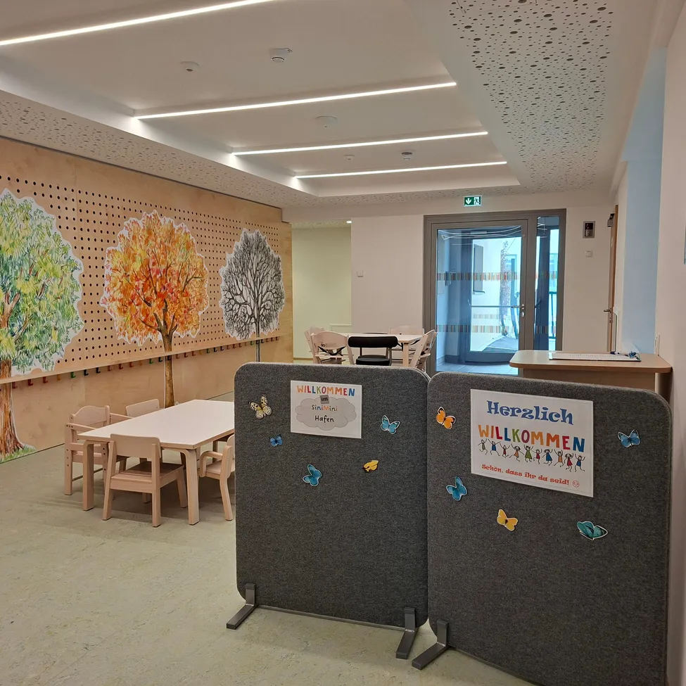 Ein Klassenzimmer mit einer großen Wandmalerei von Bäumen, einem Tisch und Stühlen sowie einem Schild, das 'Herzlich Willkommen' sagt. Der Raum ist gut beleuchtet und hat ein modernes Design.