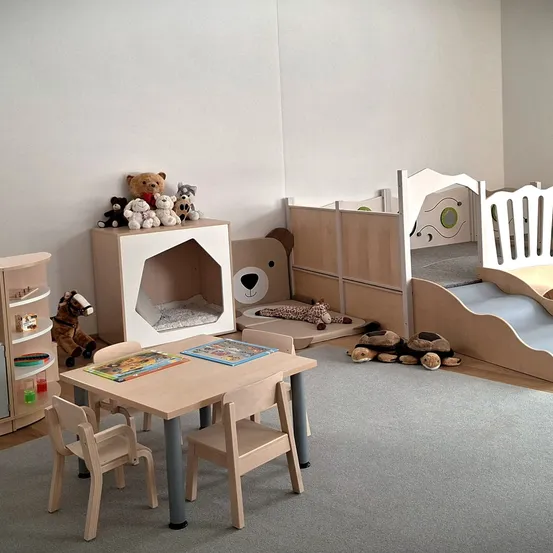 Ein Kinderzimmer mit einem Holzbett, einem Schreibtisch mit Büchern und einem Regal mit Spielzeug. Der Raum hat einen Teppichboden und weiße Wände.