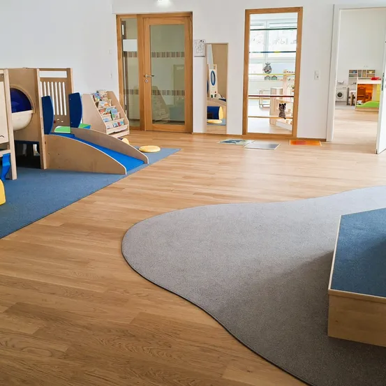 Ein Spielbereich für Kinder mit einer Holzrutsche, einem Bücherregal und einem Teppichboden. Der Raum hat Holzböden und weiße Wände.