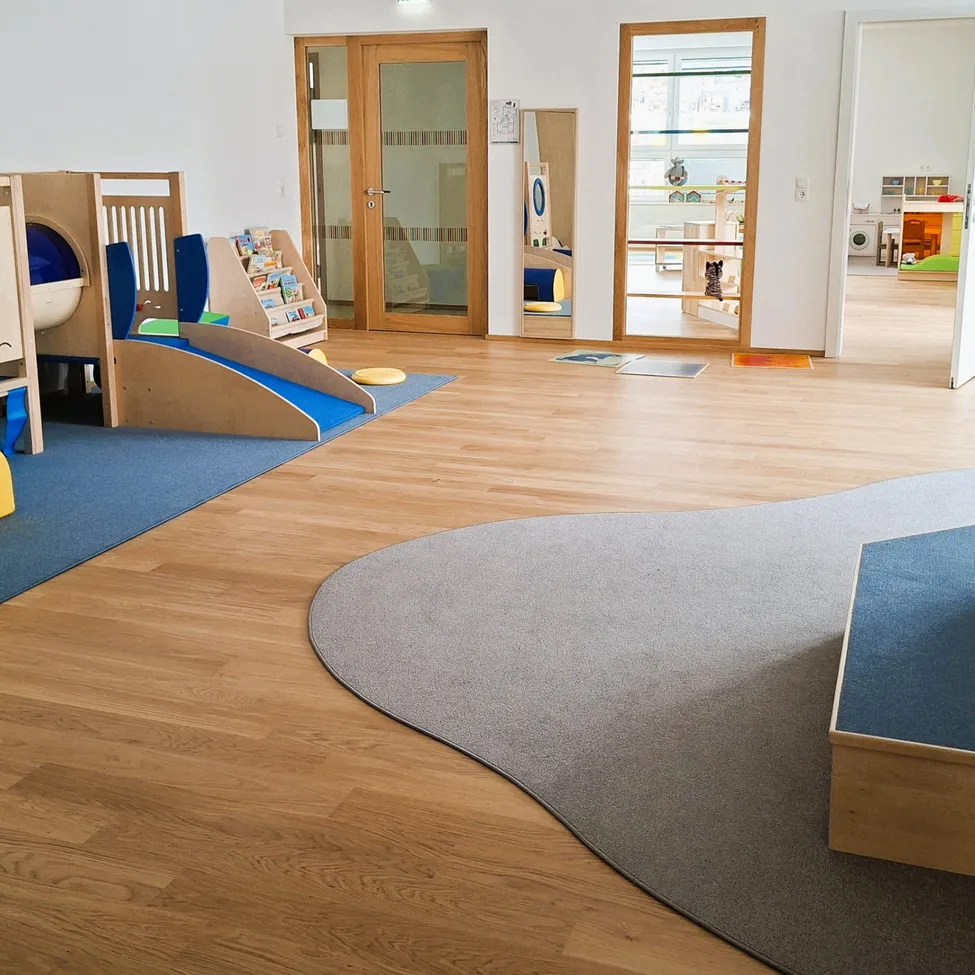 Ein Spielbereich für Kinder mit einer Holzrutsche, einem Bücherregal und einem Teppichboden. Der Raum hat Holzböden und weiße Wände.
