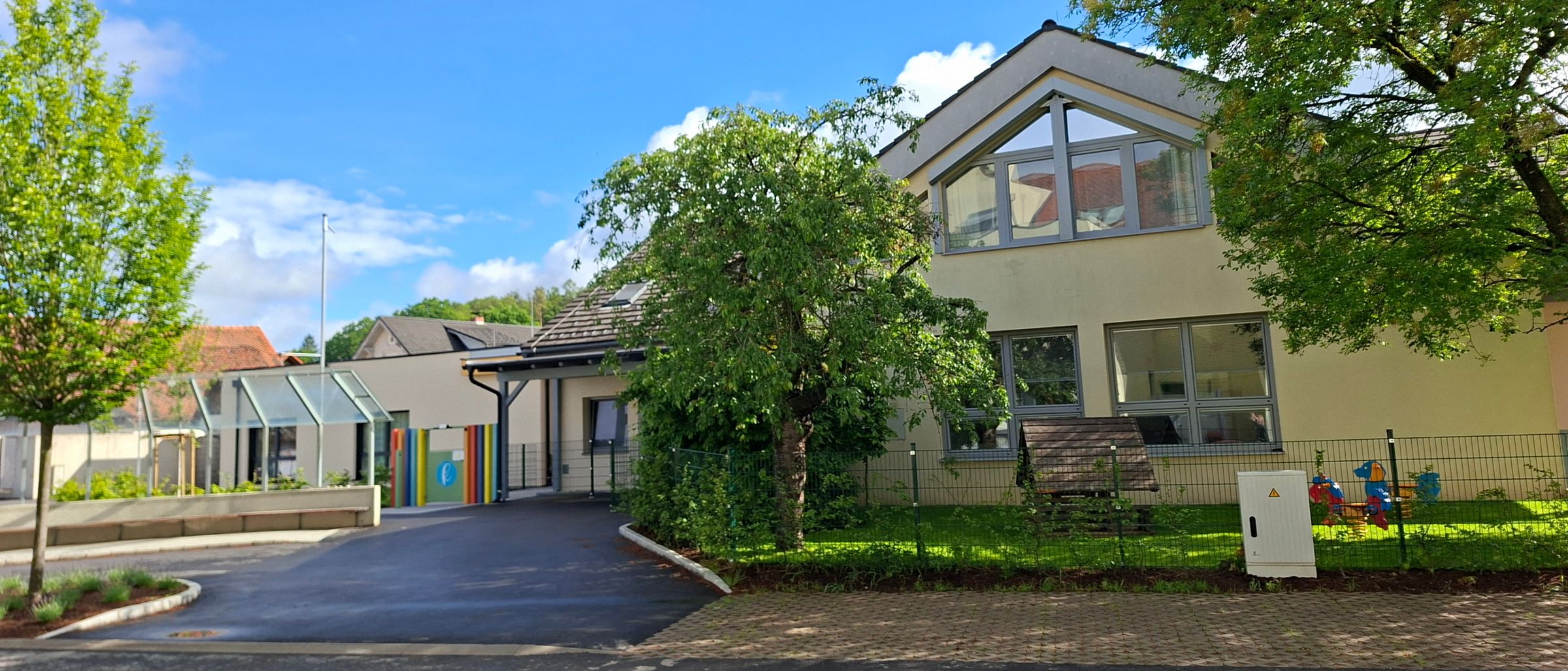 Ein modernes Haus mit großen Fenstern und einer Einfahrt, umgeben von Bäumen und einem Zaun, unter einem blauen Himmel mit Wolken.