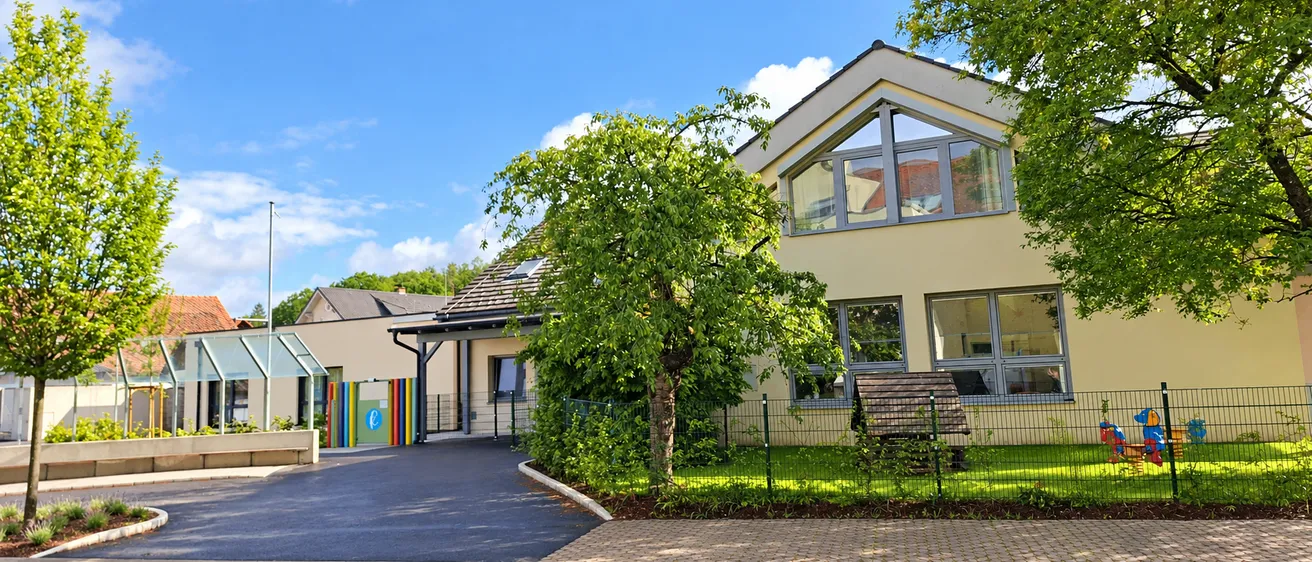 Ein gelbes Haus mit großen Fenstern und einer Einfahrt vor dem Haus, umgeben von Bäumen und einem Zaun.
