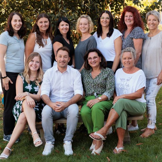 Eine Gruppe von Frauen und einem Mann posiert für ein Foto in einem Park, wobei einige auf einer Bank sitzen und andere hinter ihnen stehen. Sie alle lächeln und scheinen Kollegen oder Freunde zu sein.