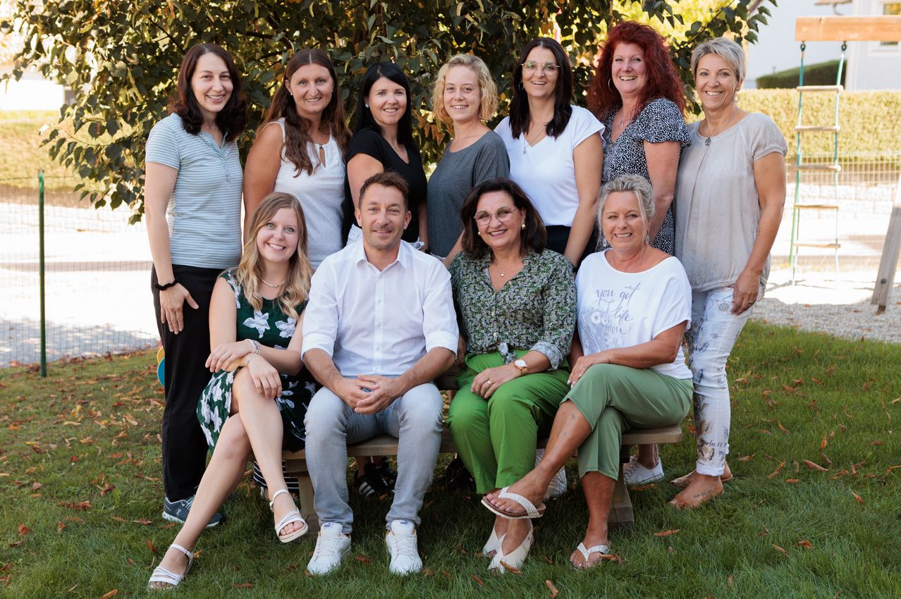 Eine Gruppe von Frauen und einem Mann posiert für ein Foto in einem Park, wobei einige auf einer Bank sitzen und andere hinter ihnen stehen. Sie alle lächeln und scheinen Kollegen oder Freunde zu sein.