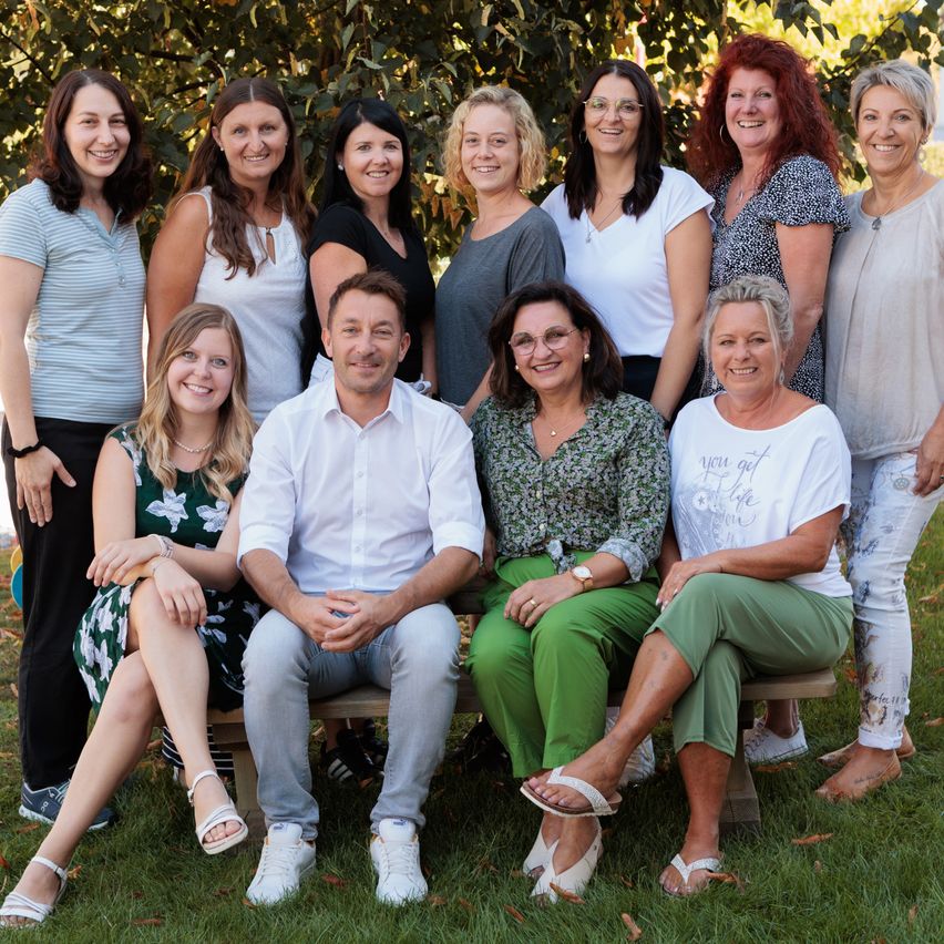Eine Gruppe von Frauen und einem Mann posiert für ein Foto in einem Park, wobei einige auf einer Bank sitzen und andere hinter ihnen stehen. Sie alle lächeln und scheinen Kollegen oder Freunde zu sein.