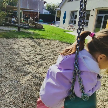 Ein kleines Mädchen in einem lila Hoodie und rosa Hosen schwingt auf einem Spielplatz auf einer Schaukel.