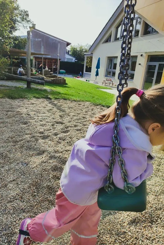 Ein kleines Mädchen in einem lila Hoodie und rosa Hosen schwingt auf einem Spielplatz auf einer Schaukel.