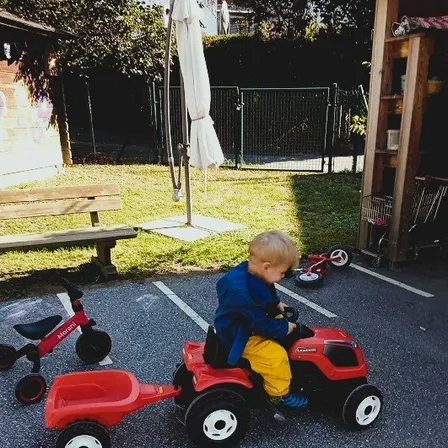 Ein kleiner Junge fährt einen roten Spielzeugtraktor mit Anhänger in einem Parkplatz. Dahinter steht ein rotes Dreirad.