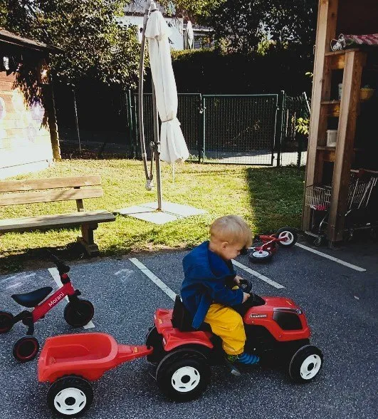 Ein kleiner Junge fährt einen roten Spielzeugtraktor mit Anhänger in einem Parkplatz. Dahinter steht ein rotes Dreirad.